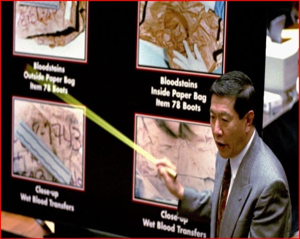 Man in a suit points to a forensic display showing bloodstain evidence in paper bags on a dark board.