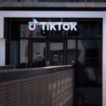 TikTok logo above glass doors of a building entrance, with a metal railing in the foreground.