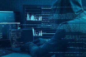 Hacker in a hoodie at a multi-monitor setup displaying code and graphs, blue-toned lighting.