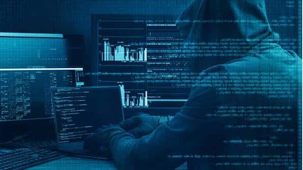 Hacker in a hoodie at a multi-monitor setup displaying code and graphs, blue-toned lighting.