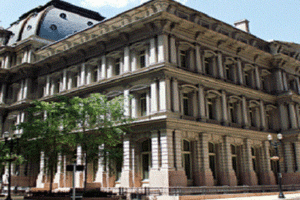 Historic neoclassical building with multiple arcs and columns along a tree-lined street on a sunny day.
