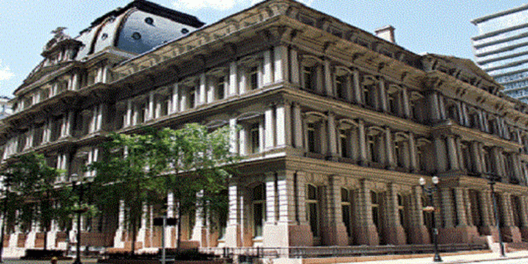 Historic neoclassical building with multiple arcs and columns along a tree-lined street on a sunny day.