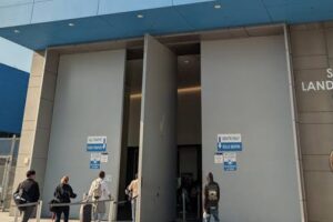 Entrance to San Ysidro Land Port of Entry, east pedestrian side, with people queued outside and a U.S. flag nearby