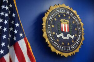 American flag beside the Department of Justice/FBI seal on a blue wall, conveying official government imagery.
