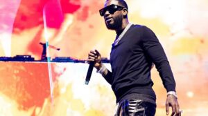 Male performer on stage with microphone, wearing sunglasses and a black outfit, against a bright orange-pink background.