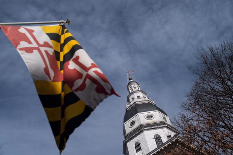Maryland state flag fluttering in the foreground with a white church steeple and bare trees against a blue sky.