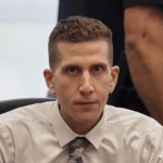 Man in a white dress shirt and patterned tie sits at a desk, looking toward the camera with a colleague standing behind him.