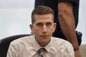 Man in a white dress shirt and patterned tie sits at a desk, looking toward the camera with a colleague standing behind him.