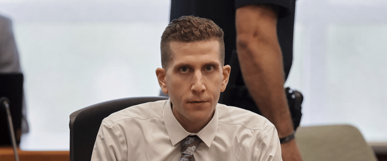 Man in a white dress shirt and patterned tie sits at a desk, looking toward the camera with a colleague standing behind him.