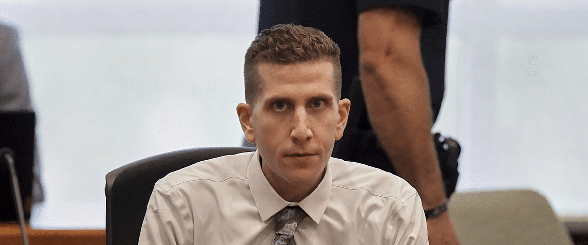 Man in a white dress shirt and patterned tie sits at a desk, looking toward the camera with a colleague standing behind him.