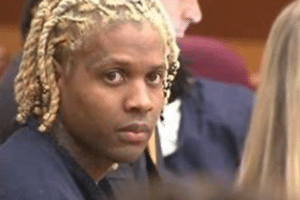 Person with blond dreadlock braids looks toward the camera in a courtroom setting, surrounded by others in formal attire.