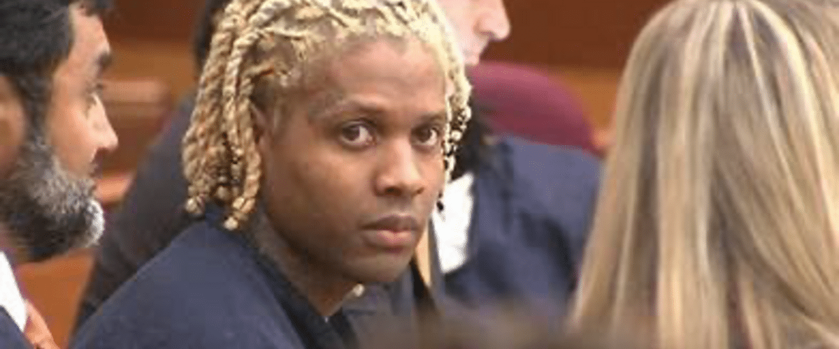Person with blond dreadlock braids looks toward the camera in a courtroom setting, surrounded by others in formal attire.