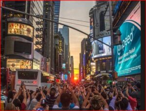 Crowd in Times Square raising phones to capture a sunset between tall neon-lit billboards and signs, with a bus in the foreground.