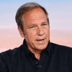 Middle-aged man in a black shirt speaks during an interview, with a blurred cityscape and a tall white obelisk in the background.