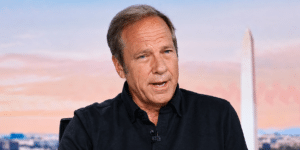 Middle-aged man in a black shirt speaks during an interview, with a blurred cityscape and a tall white obelisk in the background.