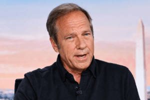 Middle-aged man in a black shirt speaks during an interview, with a blurred cityscape and a tall white obelisk in the background.
