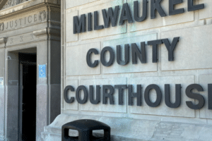 Exterior of Milwaukee County Courthouse showing a large stone sign reading MILWAUKEE COUNTY COURTHOUSE and a blue wheelchair-accessibility sign near the entrance.