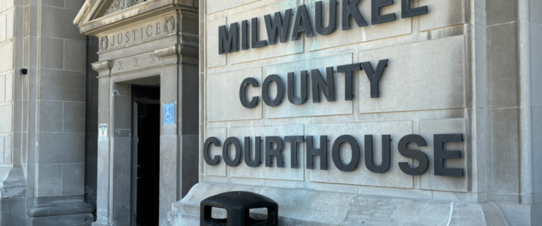 Exterior of Milwaukee County Courthouse showing a large stone sign reading MILWAUKEE COUNTY COURTHOUSE and a blue wheelchair-accessibility sign near the entrance.