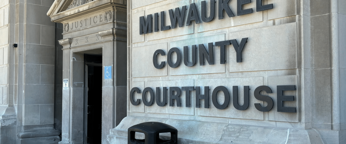 Exterior of Milwaukee County Courthouse showing a large stone sign reading MILWAUKEE COUNTY COURTHOUSE and a blue wheelchair-accessibility sign near the entrance.