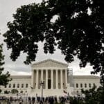 White neoclassical courthouse with tall columns, framed by leafy trees and a crowd on the steps, on a cloudy day.—Informative image