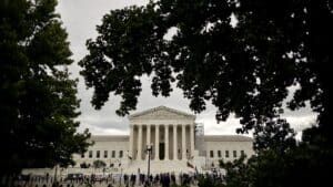White neoclassical courthouse with tall columns, framed by leafy trees and a crowd on the steps, on a cloudy day.—Informative image