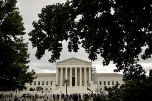 White neoclassical courthouse with tall columns, framed by leafy trees and a crowd on the steps, on a cloudy day.—Informative image