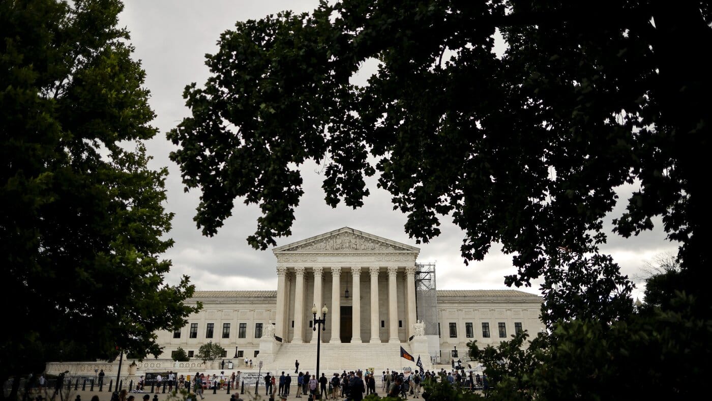 White neoclassical courthouse with tall columns, framed by leafy trees and a crowd on the steps, on a cloudy day.—Informative image