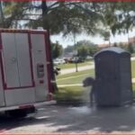 Fire truck parked on a street beside a grey portable toilet; a person is bending near it.