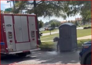 Fire truck parked on a street beside a grey portable toilet; a person is bending near it.