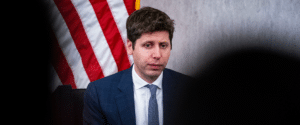 Man in a blue suit and light blue tie sits at a hearing table with an American flag behind him.