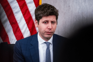 Man in a blue suit and light blue tie sits at a hearing table with an American flag behind him.