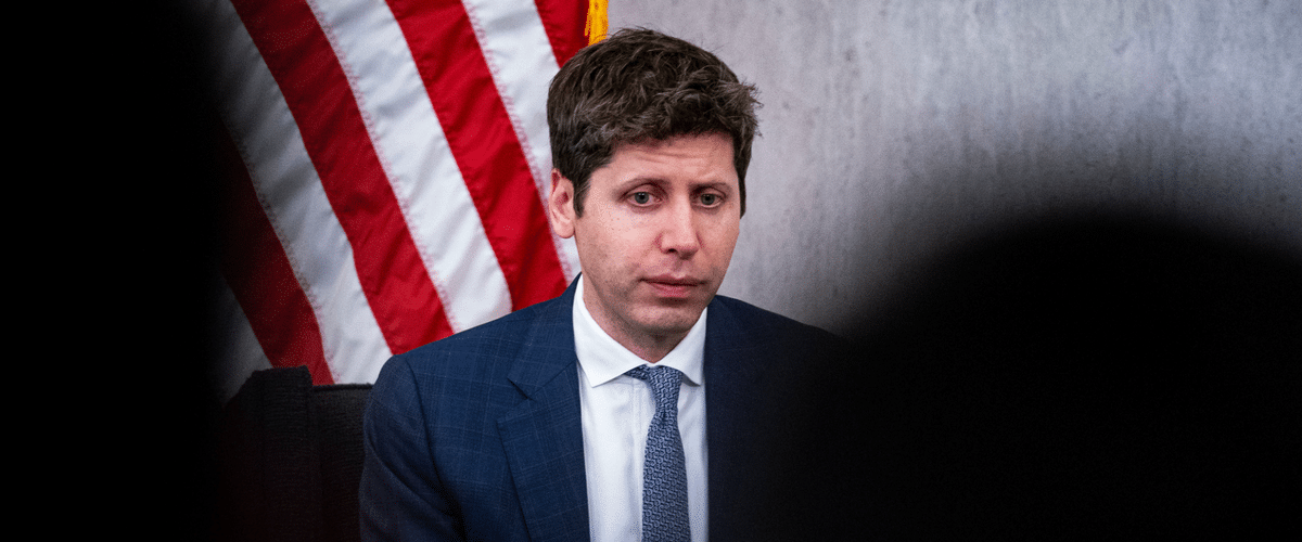 Man in a blue suit and light blue tie sits at a hearing table with an American flag behind him.