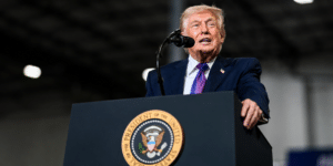 Man in a navy suit and purple tie speaks at a podium with a circular eagle seal in front of him.