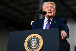 Man in a navy suit and purple tie speaks at a podium with a circular eagle seal in front of him.