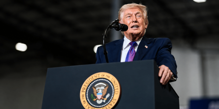Man in a navy suit and purple tie speaks at a podium with a circular eagle seal in front of him.