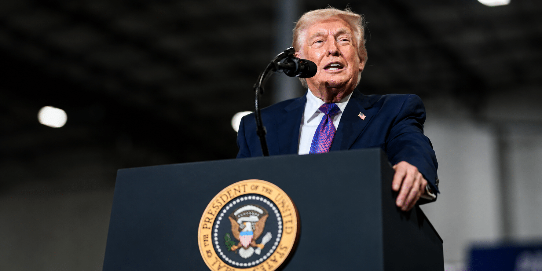 Man in a navy suit and purple tie speaks at a podium with a circular eagle seal in front of him.