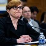 Panelist with short blond hair and glasses sits at a table with a microphone, hands folded in front, in a conference room.