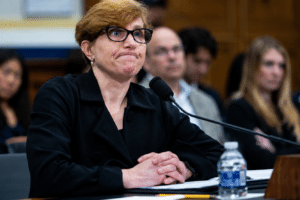 Panelist with short blond hair and glasses sits at a table with a microphone, hands folded in front, in a conference room.
