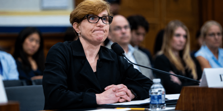 Panelist with short blond hair and glasses sits at a table with a microphone, hands folded in front, in a conference room.