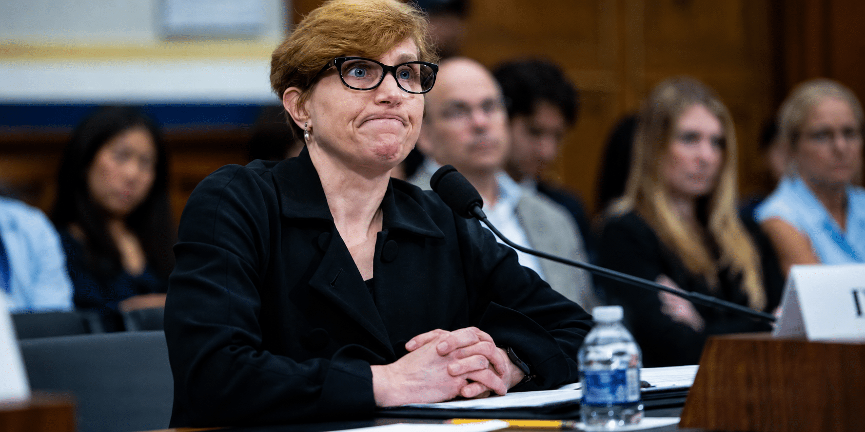Panelist with short blond hair and glasses sits at a table with a microphone, hands folded in front, in a conference room.