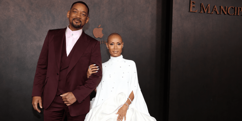 Will Smith and Jada Pinkett Smith pose arm-in-arm on a dark backdrop at an Emancipation premiere in formal attire (burgundy suit and white gown).