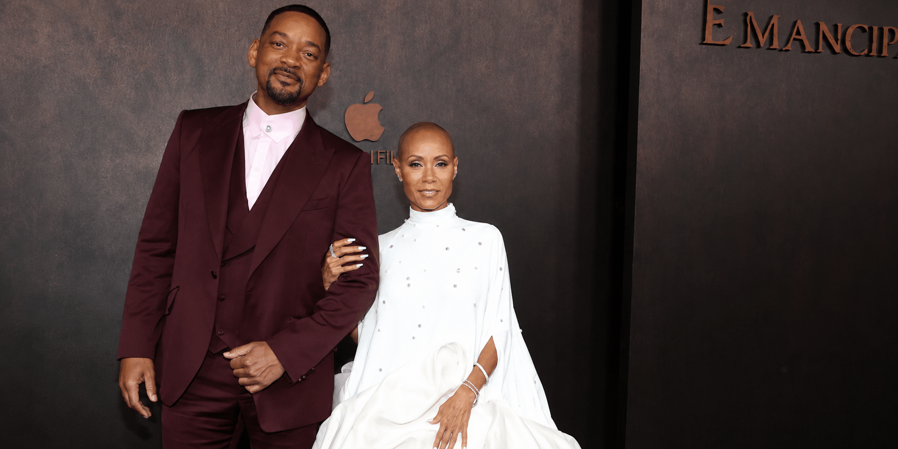 Will Smith and Jada Pinkett Smith pose arm-in-arm on a dark backdrop at an Emancipation premiere in formal attire (burgundy suit and white gown).