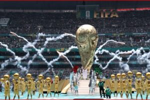 World Cup trophy on a pedestal surrounded by gold-clad performers in a stadium with fireworks and cheering crowds