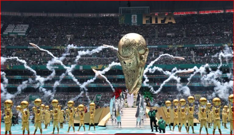 World Cup trophy on a pedestal surrounded by gold-clad performers in a stadium with fireworks and cheering crowds