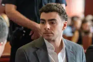 Man in a gray suit with a white shirt sits in a barbershop, a barber's arm visible behind him.