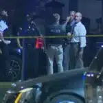 Nighttime police scene with officers behind yellow caution tape at a cordoned street area, a police SUV in foreground and a group of investigators in discussion.