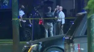 Nighttime police scene with officers behind yellow caution tape at a cordoned street area, a police SUV in foreground and a group of investigators in discussion.