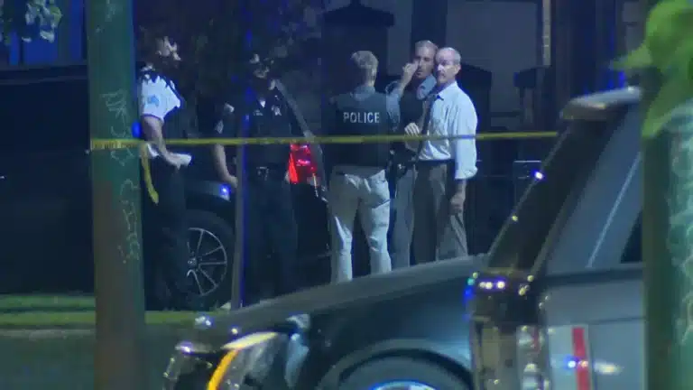 Nighttime police scene with officers behind yellow caution tape at a cordoned street area, a police SUV in foreground and a group of investigators in discussion.