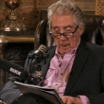 Older man in a blazer reads from a notebook while speaking into a mounted microphone in a cozy, wood-paneled studio set with a leather chair behind him.