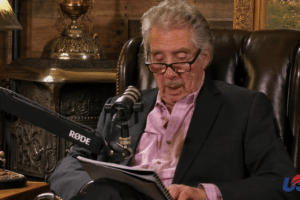 Older man in a blazer reads from a notebook while speaking into a mounted microphone in a cozy, wood-paneled studio set with a leather chair behind him.